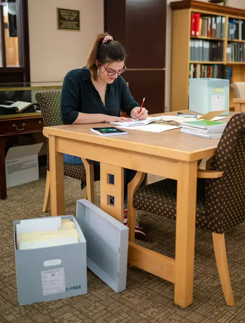 Anastasija Mladenovska sits at a table in the reading room of the Walter Havighurst Special Collections and Archives in King Library. She is looking down at a small open book on the table and holding a pencil over a notebook. There are two open archival file boxes containing books and documents on the table and on the floor around her, and two small stacks of books and documents also sit on the table near her.